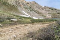 a dirt path in front of a grassy mountain side near water and snow mountains with a trail going down it