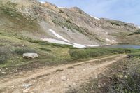 a dirt path in front of a grassy mountain side near water and snow mountains with a trail going down it