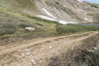 a dirt path in front of a grassy mountain side near water and snow mountains with a trail going down it