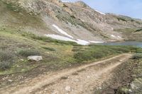 a dirt path in front of a grassy mountain side near water and snow mountains with a trail going down it