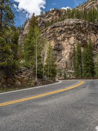 the curved road curves in front of a mountain area with pine trees behind it,