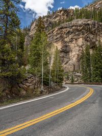 the curved road curves in front of a mountain area with pine trees behind it,