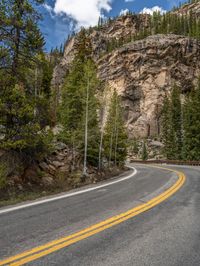 the curved road curves in front of a mountain area with pine trees behind it,