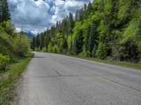 a street leads to a narrow forest with mountains in the background with clouds overhead on an empty mountain road