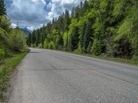 a street leads to a narrow forest with mountains in the background with clouds overhead on an empty mountain road