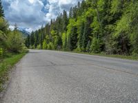 a street leads to a narrow forest with mountains in the background with clouds overhead on an empty mountain road