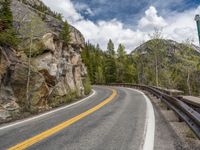 the curved road curves in front of a mountain area with pine trees behind it,