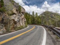 the curved road curves in front of a mountain area with pine trees behind it,