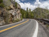 the curved road curves in front of a mountain area with pine trees behind it,