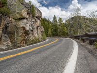 the curved road curves in front of a mountain area with pine trees behind it,