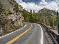 the curved road curves in front of a mountain area with pine trees behind it,