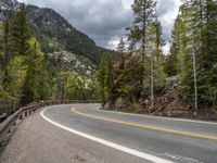 the curved road curves in front of a mountain area with pine trees behind it,