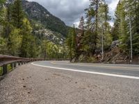 the curved road curves in front of a mountain area with pine trees behind it,