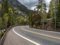 the curved road curves in front of a mountain area with pine trees behind it,