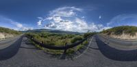 a fish eye view of the curved road next to mountains with low clouds overhead and a sky with a few white clouds