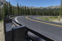 an empty asphalt road near pine covered mountains and a forest filled with evergreen trees under a blue sky