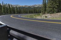 an empty asphalt road near pine covered mountains and a forest filled with evergreen trees under a blue sky