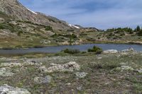 a small lake sits at the edge of a slope and is surrounded by shrubs, rocks, and snow