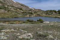 a small lake sits at the edge of a slope and is surrounded by shrubs, rocks, and snow