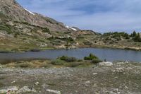 a small lake sits at the edge of a slope and is surrounded by shrubs, rocks, and snow