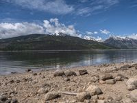 a lake with rocks on the shore and mountains in the background along side it and snow capped hills