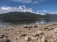 a lake with rocks on the shore and mountains in the background along side it and snow capped hills