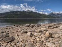 a lake with rocks on the shore and mountains in the background along side it and snow capped hills