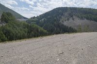 a dirt and gravel road with a mountain range in the background near a forest and valley