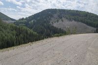 a dirt and gravel road with a mountain range in the background near a forest and valley