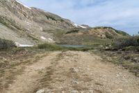 a dirt trail winds through a mountain area near a lake with a blue sky and white clouds