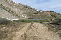 a dirt trail winds through a mountain area near a lake with a blue sky and white clouds