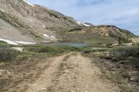 a dirt trail winds through a mountain area near a lake with a blue sky and white clouds