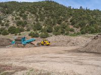 two yellow bulldozers work on sand and gravel in a mountain valley while another one looks at it