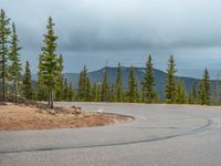 there is a black sign that is on a road with mountains in the background, on top of a hill near trees