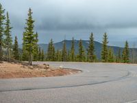 there is a black sign that is on a road with mountains in the background, on top of a hill near trees