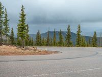 there is a black sign that is on a road with mountains in the background, on top of a hill near trees