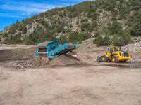 two yellow bulldozers work on sand and gravel in a mountain valley while another one looks at it