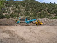 two yellow bulldozers work on sand and gravel in a mountain valley while another one looks at it