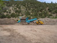 two yellow bulldozers work on sand and gravel in a mountain valley while another one looks at it