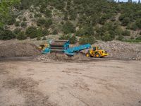 two yellow bulldozers work on sand and gravel in a mountain valley while another one looks at it