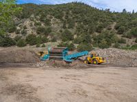 two yellow bulldozers work on sand and gravel in a mountain valley while another one looks at it