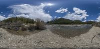 a 360 view is of a river next to some mountains with trees on each side and clouds in the blue sky above it