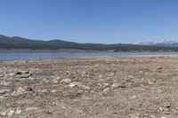 an elephant standing on the bank of a lake near mountains and rocks below it, with a small mountain range in the distance