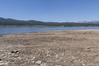 an elephant standing on the bank of a lake near mountains and rocks below it, with a small mountain range in the distance