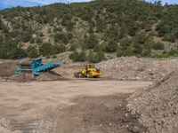 two yellow bulldozers work on sand and gravel in a mountain valley while another one looks at it