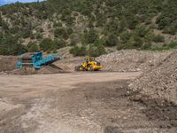 two yellow bulldozers work on sand and gravel in a mountain valley while another one looks at it