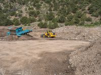 two yellow bulldozers work on sand and gravel in a mountain valley while another one looks at it