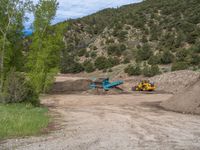 two yellow bulldozers work on sand and gravel in a mountain valley while another one looks at it