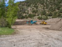 two yellow bulldozers work on sand and gravel in a mountain valley while another one looks at it