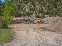 two yellow bulldozers work on sand and gravel in a mountain valley while another one looks at it